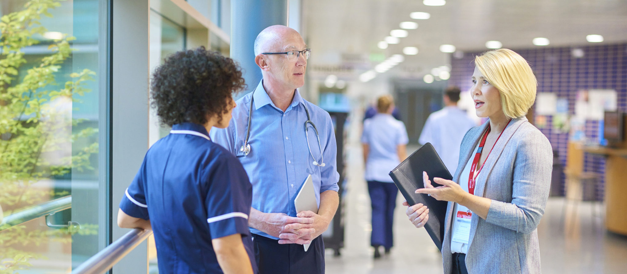 a business woman chats to a doctor and senior staff nurse in a busy hospital corridor